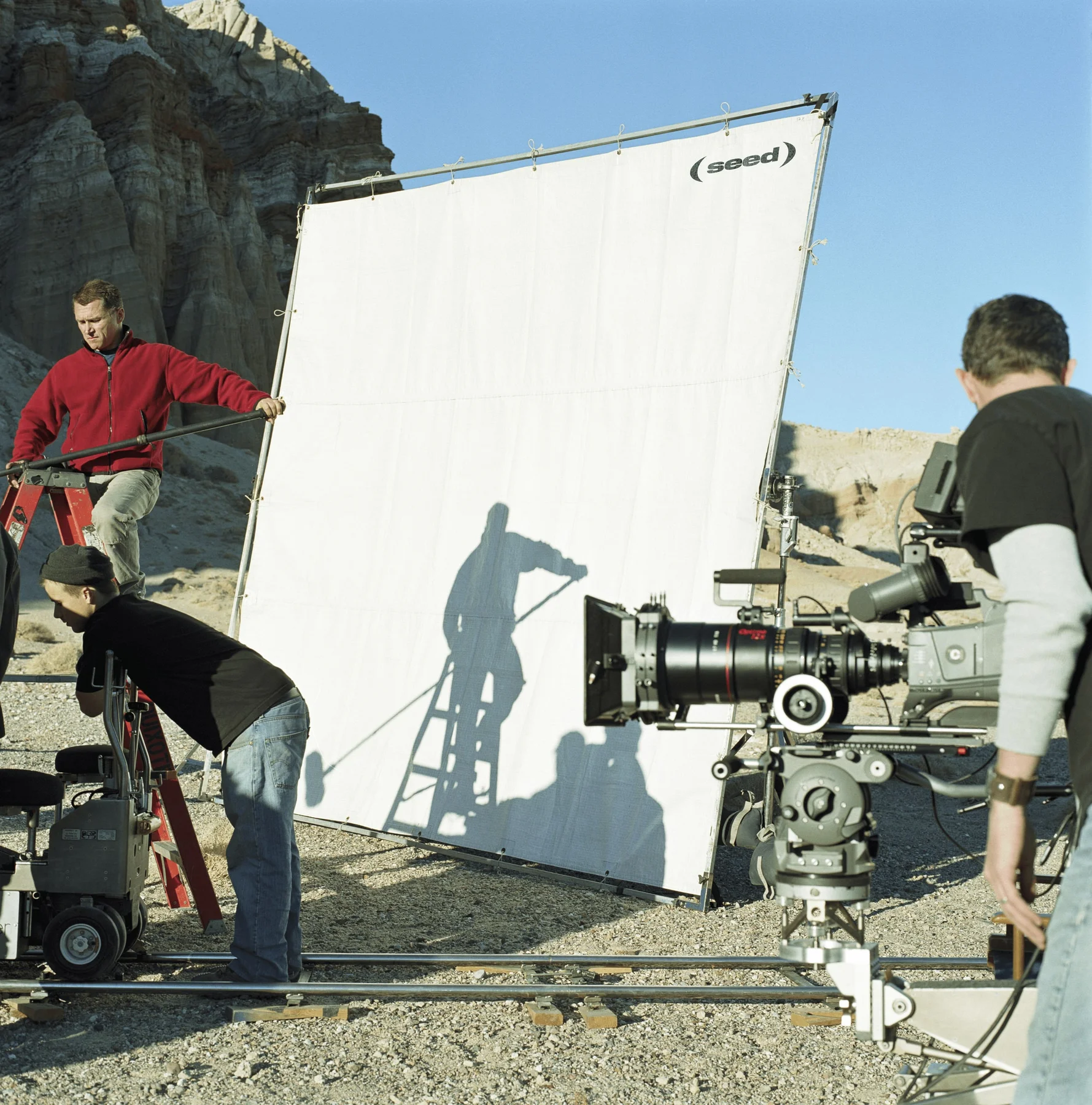 Film crew working outdoors beside a large Seed-branded reflector.