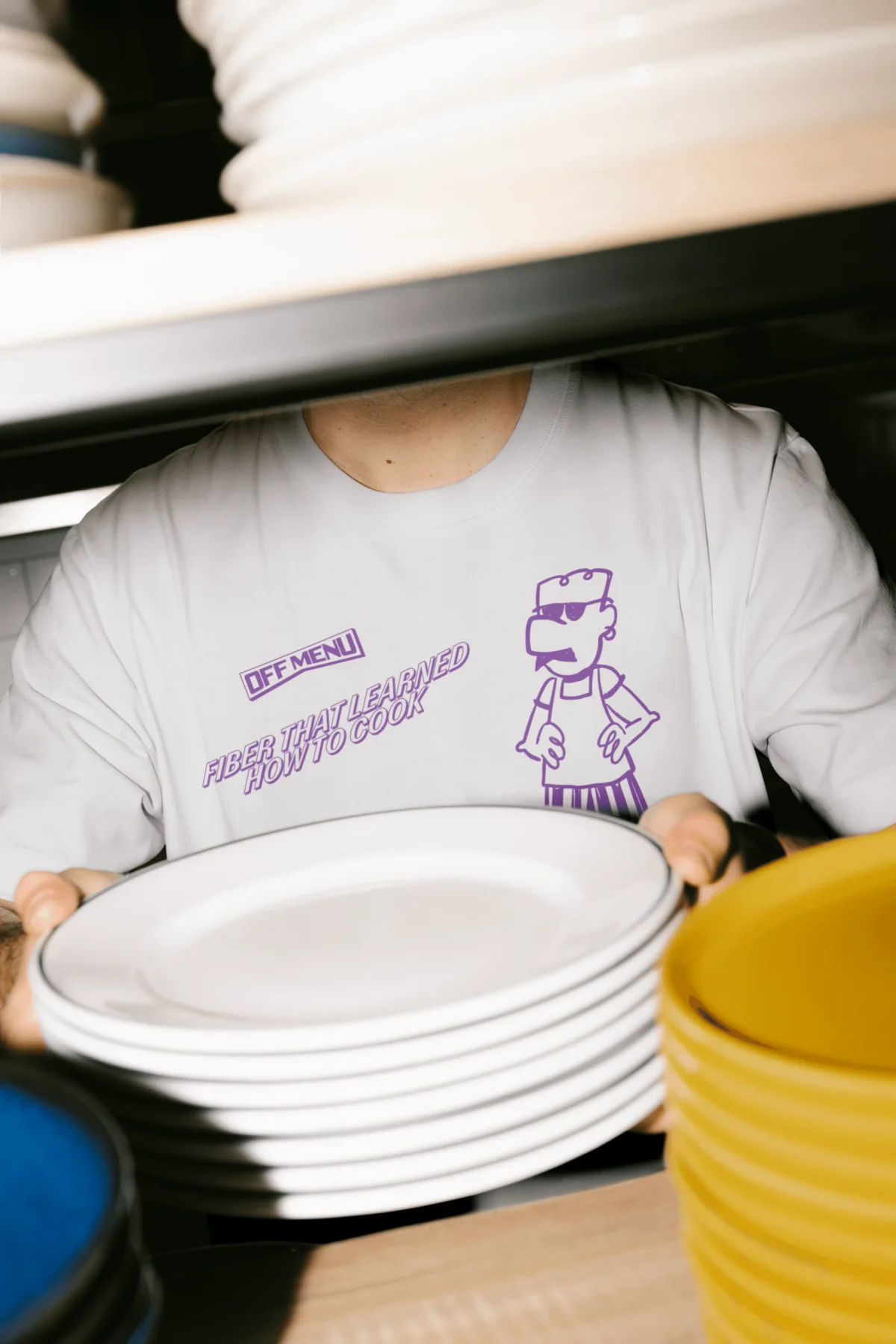 Person in an Off Menu illustrated shirt behind stacked plates.