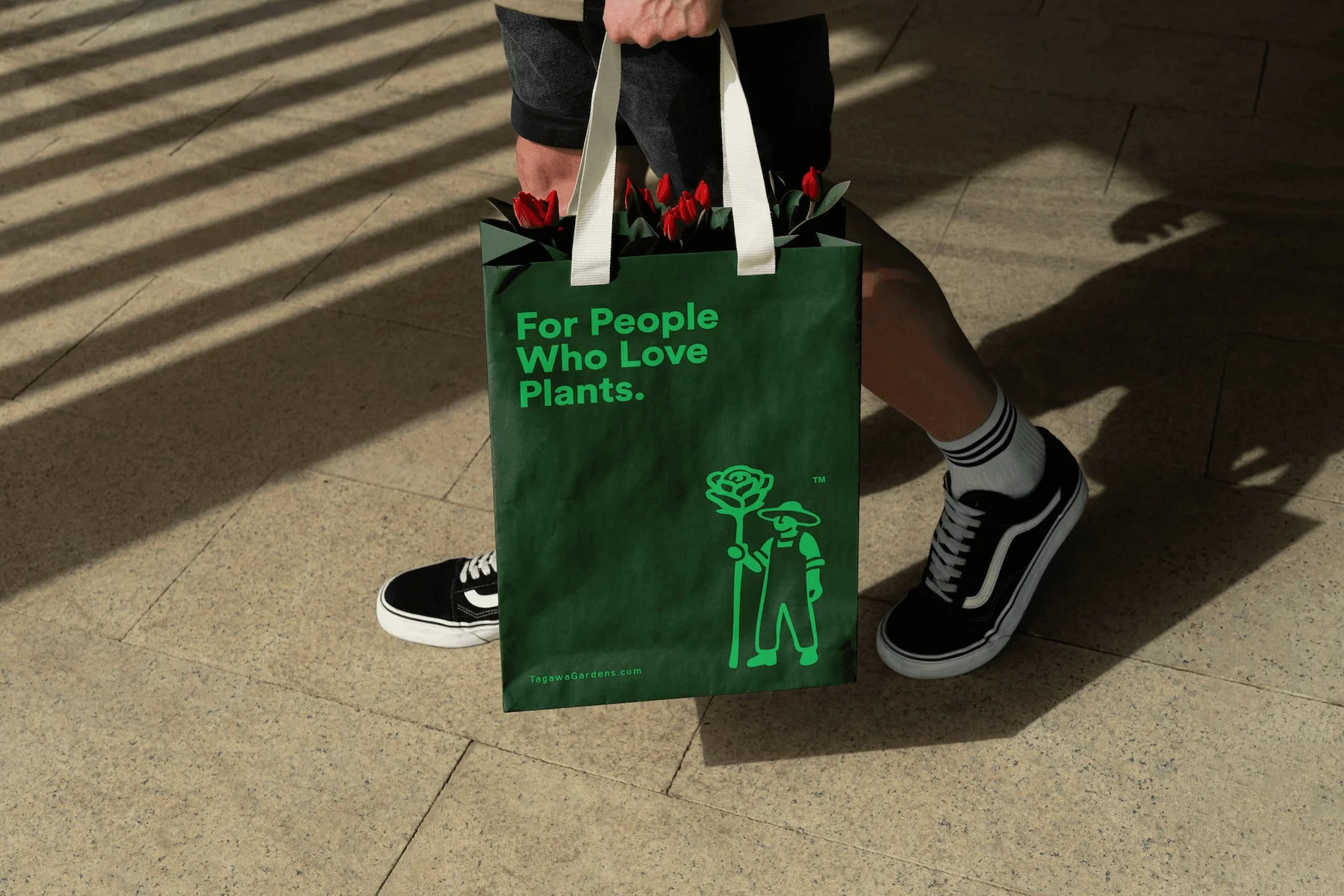 Person carrying a green Tagawa Gardens bag with a gardener illustration.