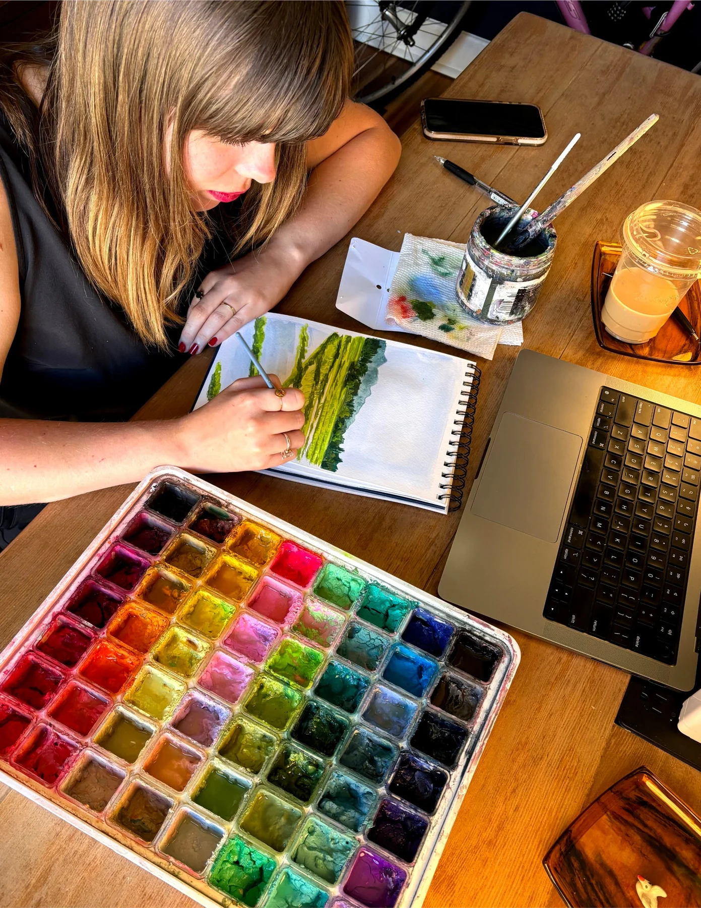 Artist painting a landscape beside a watercolor palette.