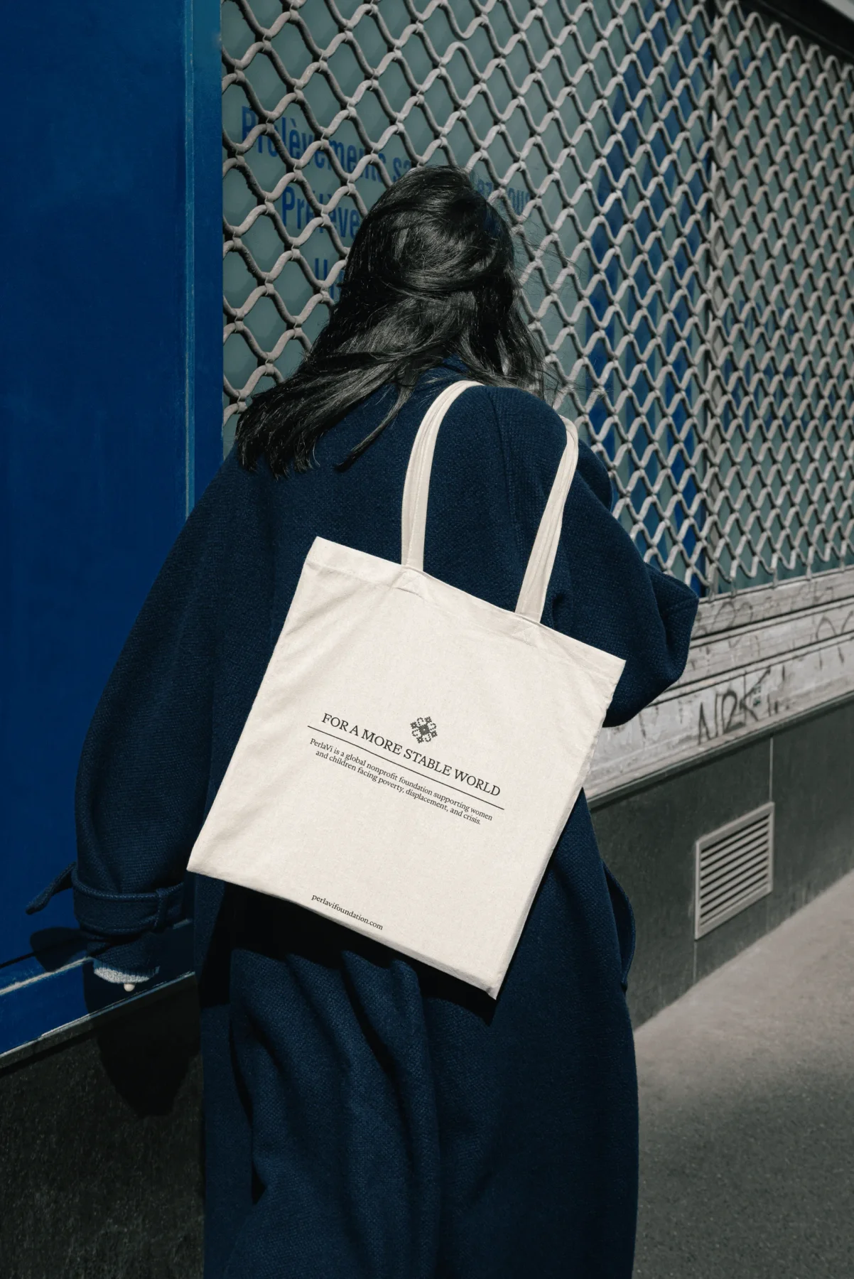 Person carrying a cream PerlaVi tote bag beside a chain-link fence.